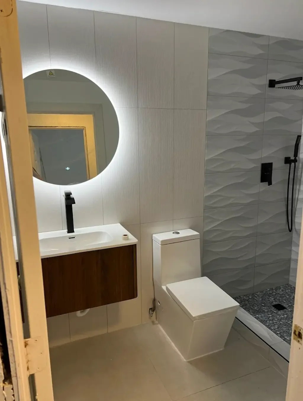 Bathroom with white wave-texture shower tile, black rain shower fixtures, pebble shower floor, backlit round mirror and wood floating vanity