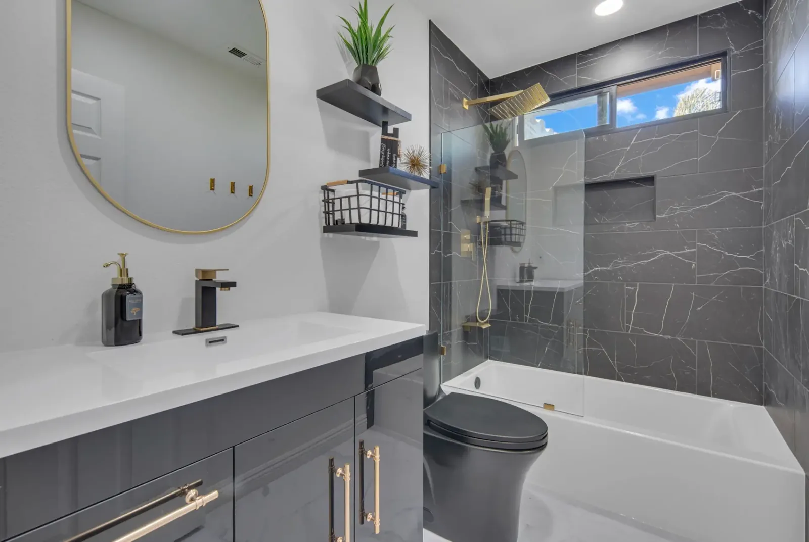 Bathroom with black marble-look tile tub and shower, frameless glass panel, gold rain shower head, recessed niche and gray gloss vanity with gold pulls