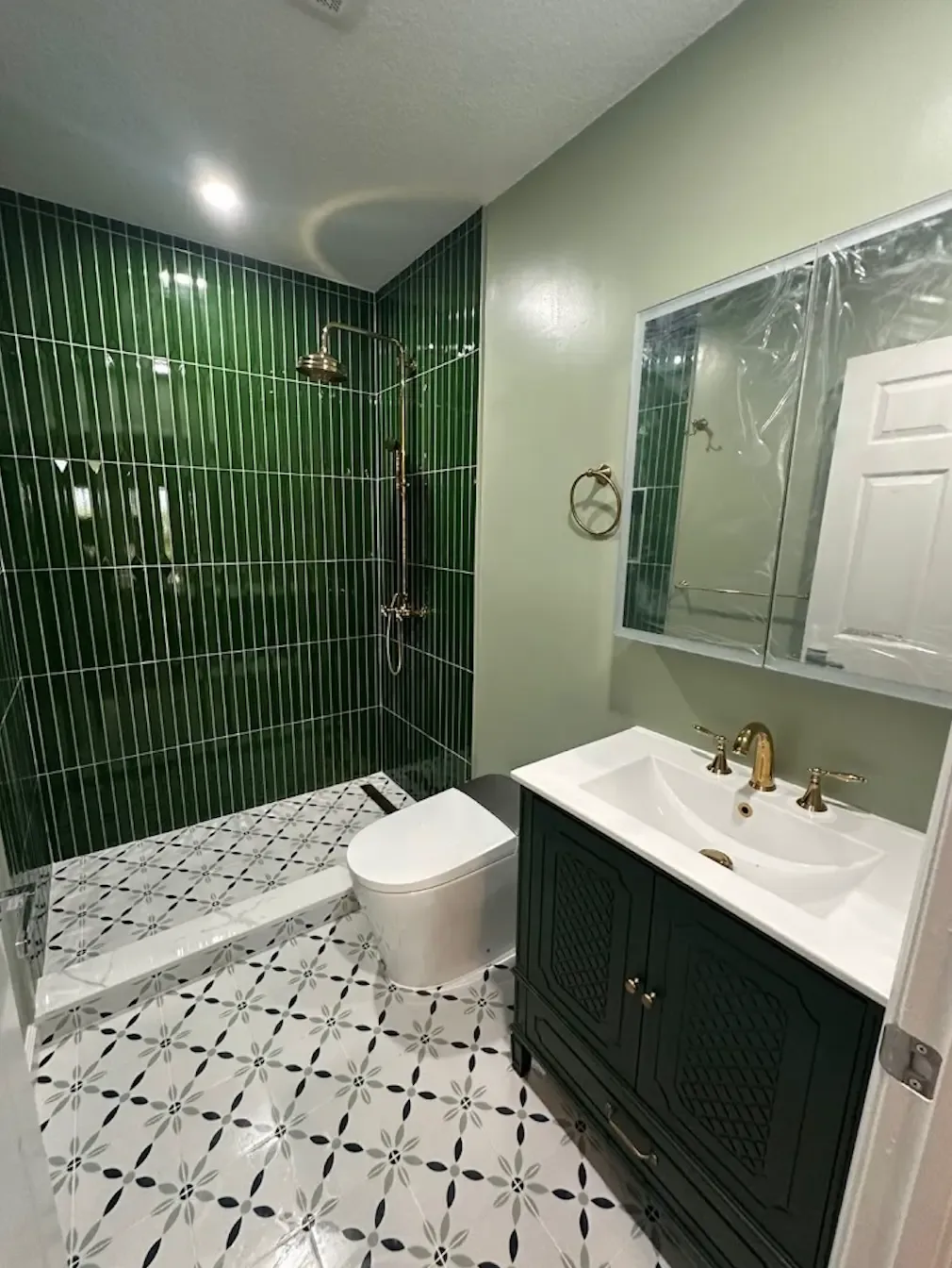 Bathroom with green vertical stacked tile walk-in shower, gold exposed shower set, patterned floor tile and dark green vanity with gold faucet