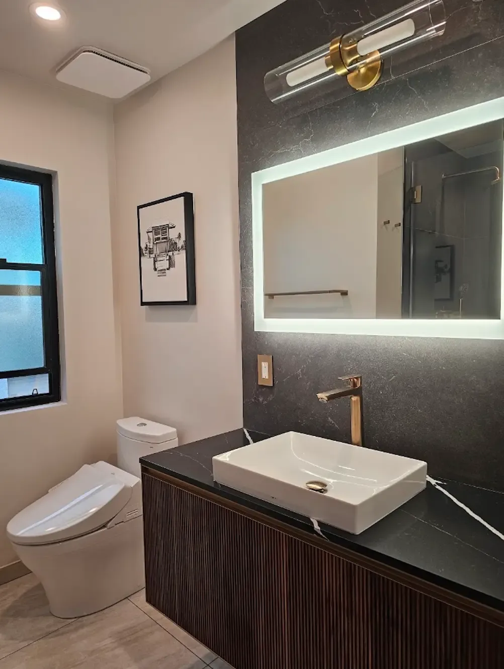 Powder bathroom with floating fluted wood vanity, black veined stone top and wall, white vessel sink, gold faucet and backlit mirror