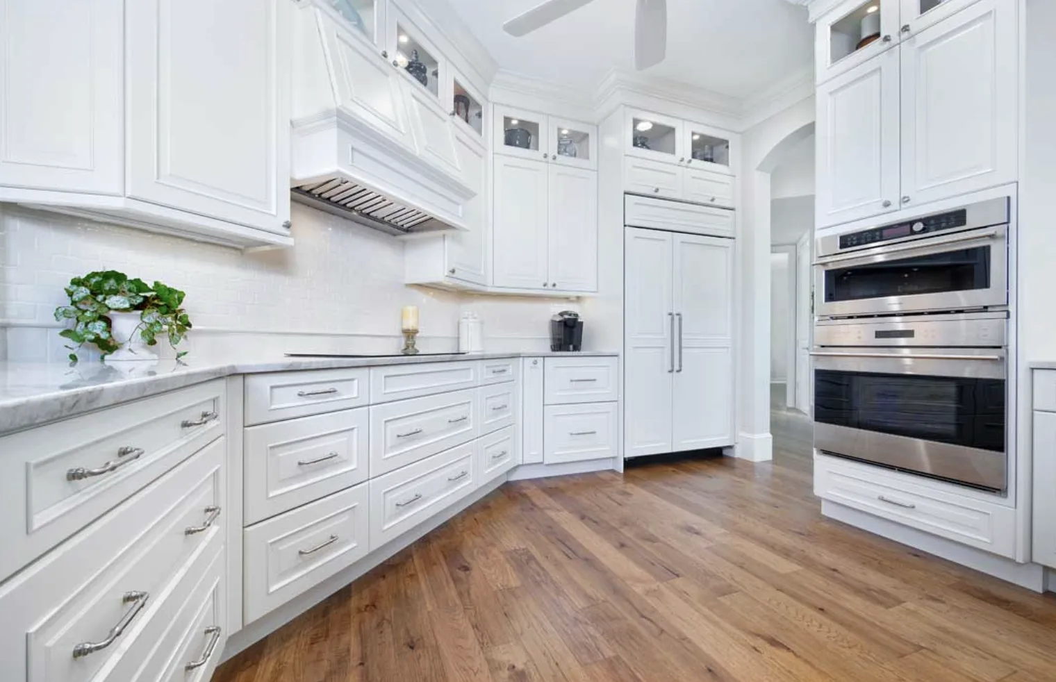White raised-panel kitchen cabinetry with glass-front uppers, paneled refrigerator, wall ovens and wood plank floor