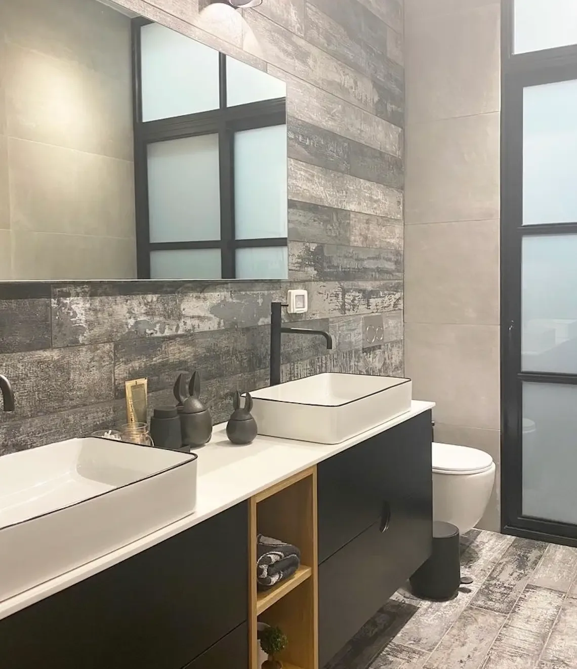Bathroom with black floating double vanity, white vessel sinks, matte black faucets, open wood shelf and distressed wood-look wall and floor tile