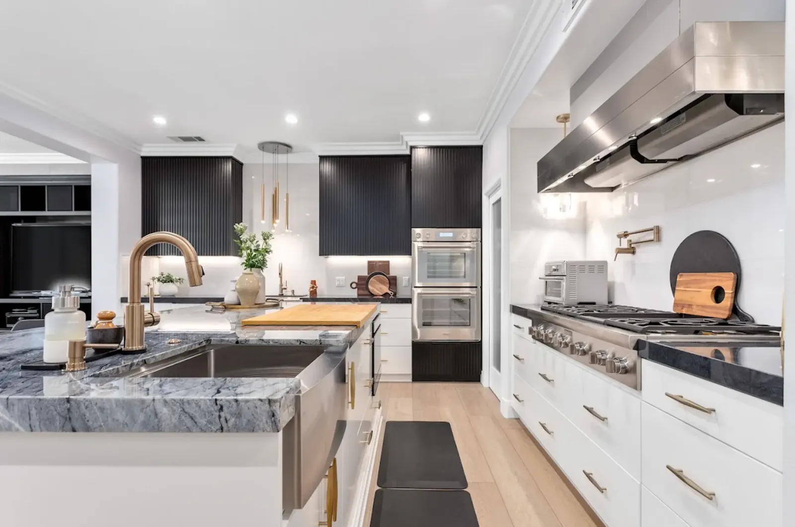 White kitchen with black fluted upper cabinets, gray veined stone island with farmhouse sink, brass faucet and pendants, double wall ovens and range hood