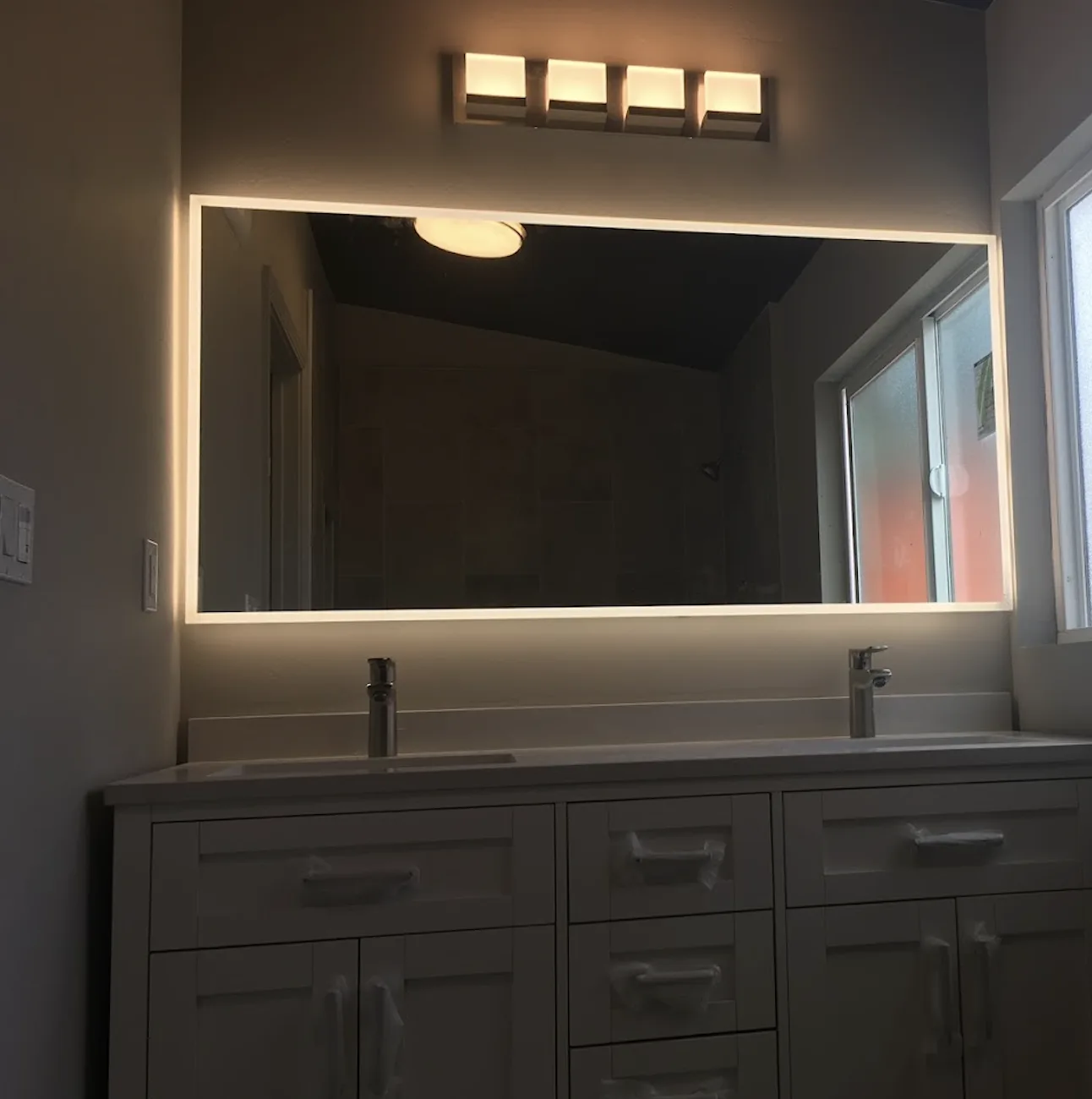 White shaker double vanity with protective wrap still on the pulls, backlit rectangular mirror and four-light vanity fixture